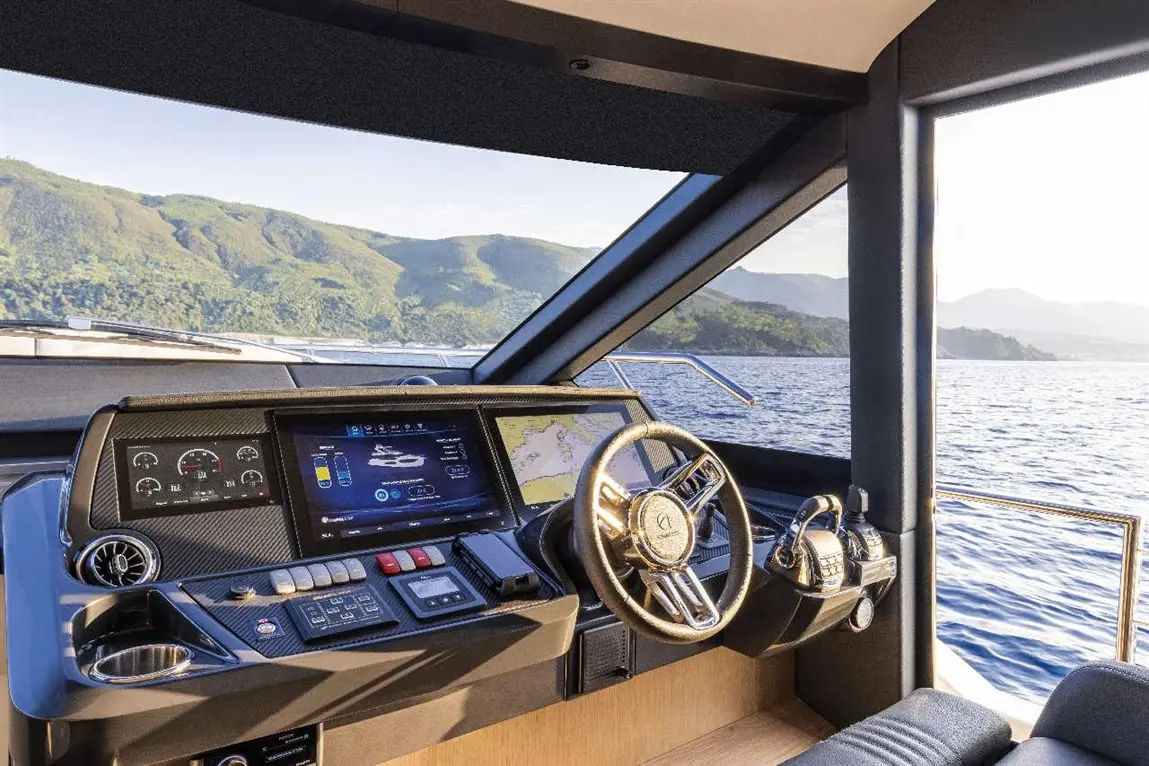 Modern yacht cockpit showcasing a steering wheel, navigation screens, and panoramic views of mountains and water.